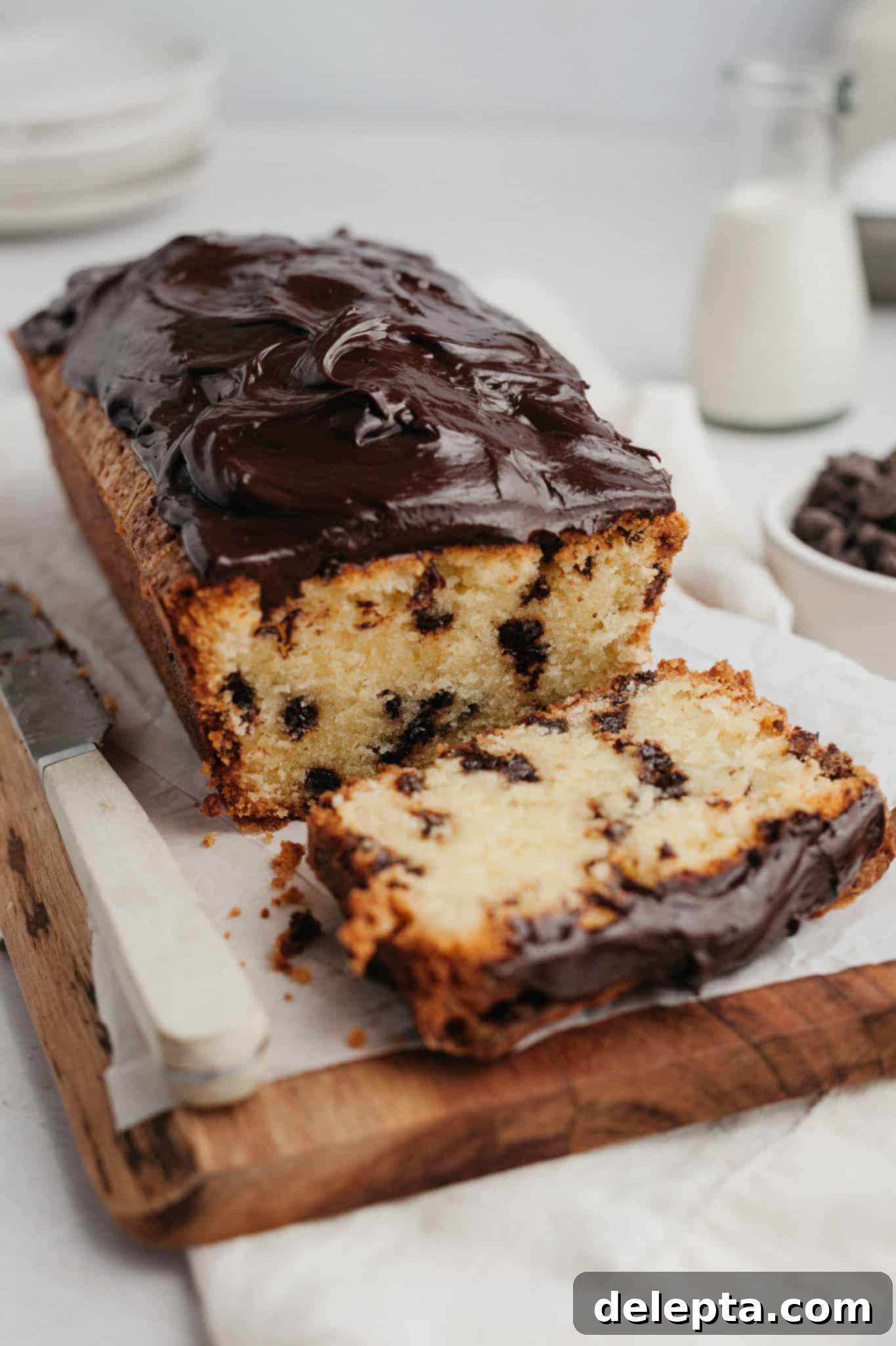 The Ultimate Chocolate Chip Loaf 2 A perfectly baked chocolate chip loaf cake, generously covered in rich chocolate ganache, sits on a rustic wooden board. One inviting slice has been cut, revealing the moist interior and abundance of mini chocolate chips.