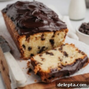 The Ultimate Chocolate Chip Loaf 8 A chocolate chip loaf cake covered in chocolate ganache on a wooden board, one slice has been cut.