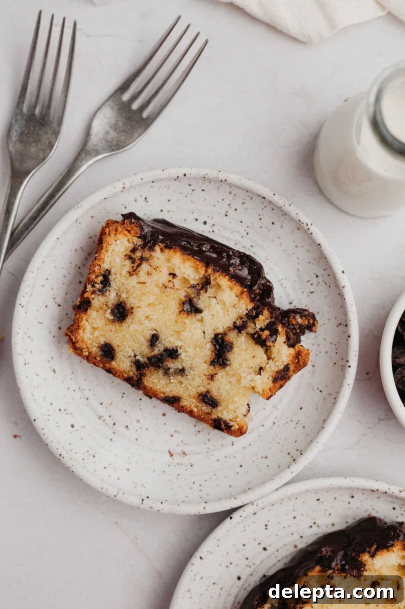 The Ultimate Chocolate Chip Loaf 7 A delicious slice of moist chocolate chip pound cake, topped with a beautiful chocolate ganache, rests on a small, speckled ceramic plate.