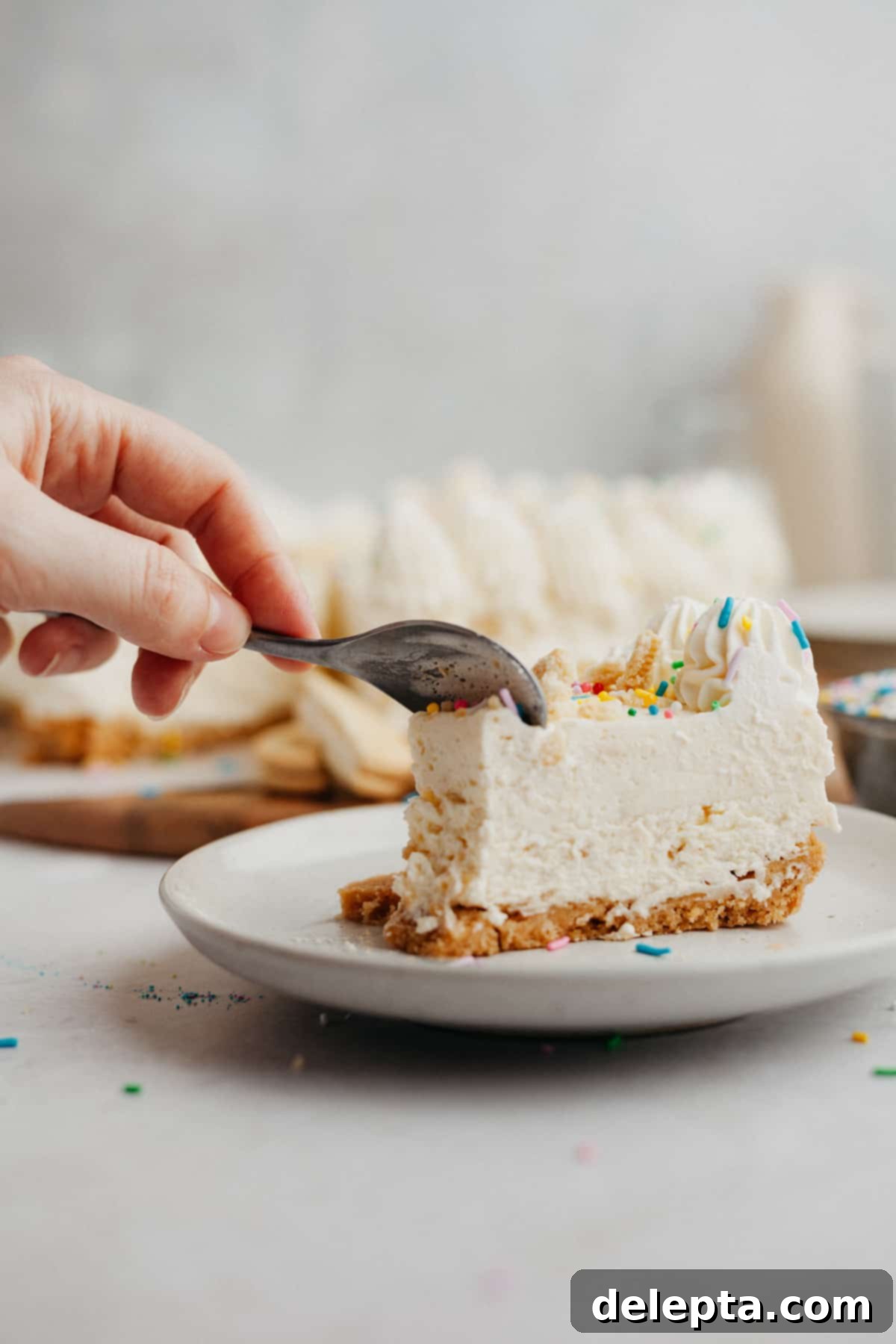 Blonde Oreo Cheesecake 12 A delightful slice of no-bake Golden Oreo cheesecake, topped with rainbow sprinkles, rests on a small beige plate. A hand gently cuts into the slice with a small silver spoon.