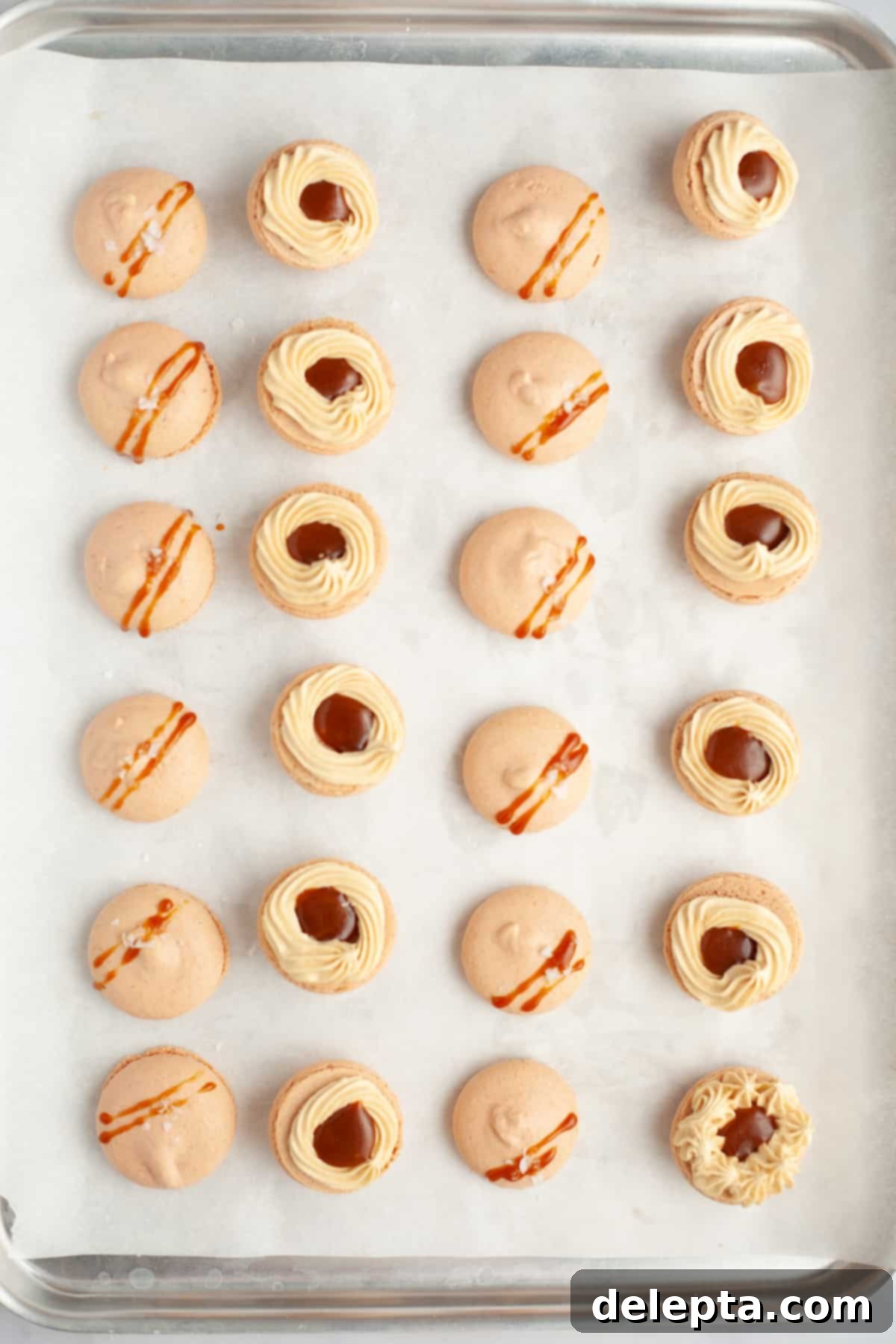 Sea Salt Caramel Macarons 18 A parchment-lined tray displaying assembled salted caramel macarons. Half are filled with buttercream and caramel, and some have caramel drizzled on top.