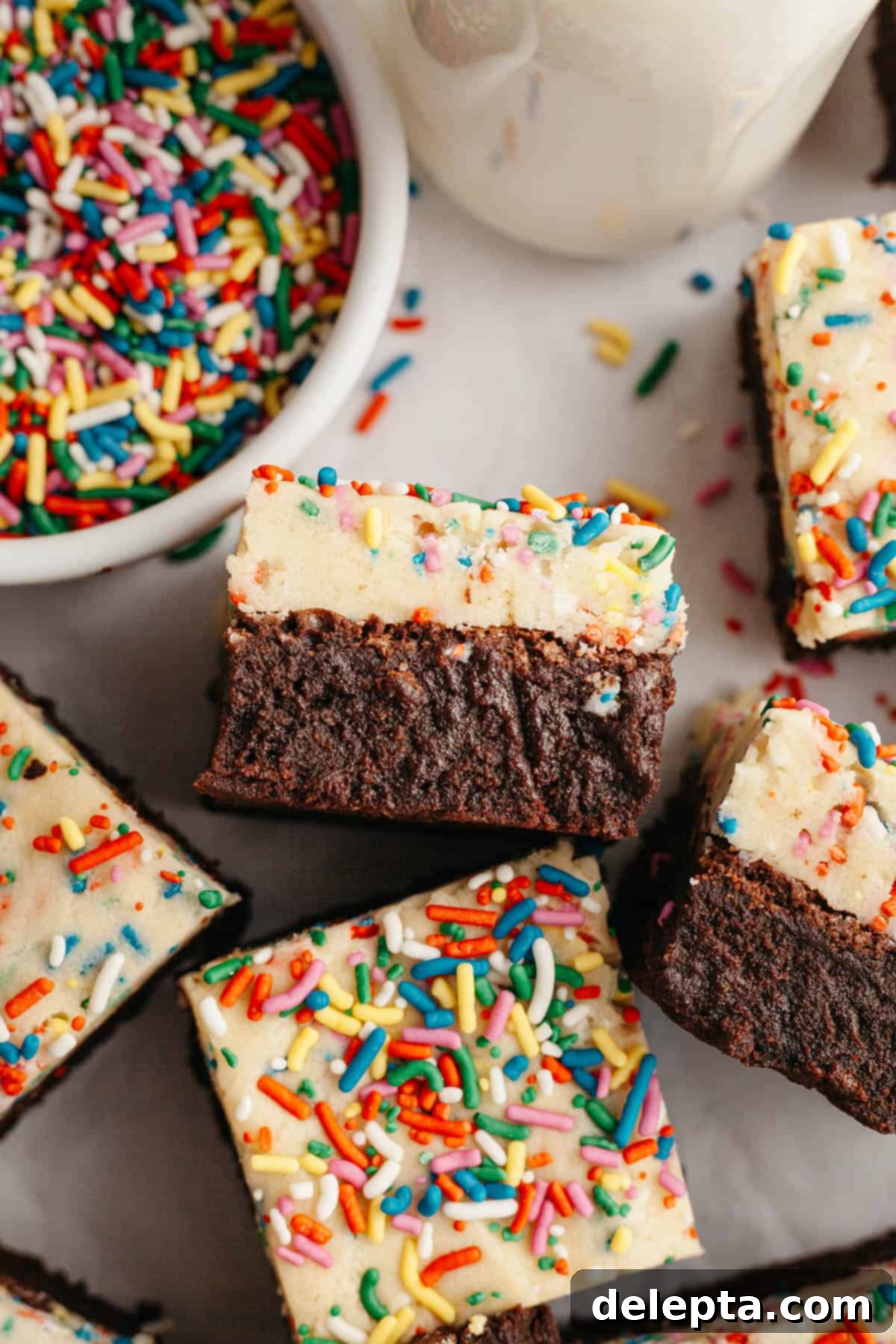 Funfetti Dream Brownies 2 Brownies with an edible cookie dough layer with rainbow sprinkles on parchment paper.