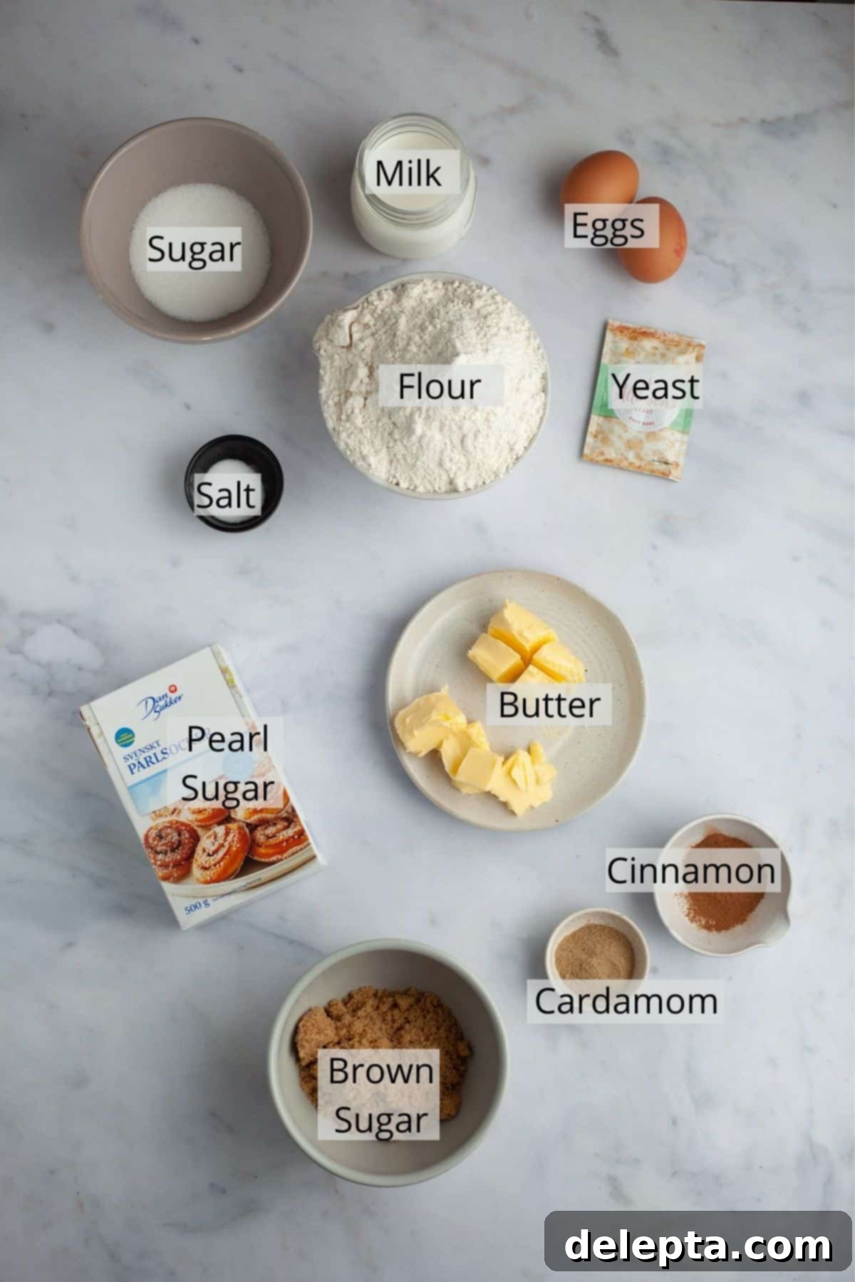 Pillowy Swedish Cinnamon Swirls 3 A flat lay photograph showcasing all the ingredients needed to make Swedish cinnamon rolls, including flour, sugar, butter, milk, eggs, yeast, ground cinnamon, cardamom, and pearl sugar.