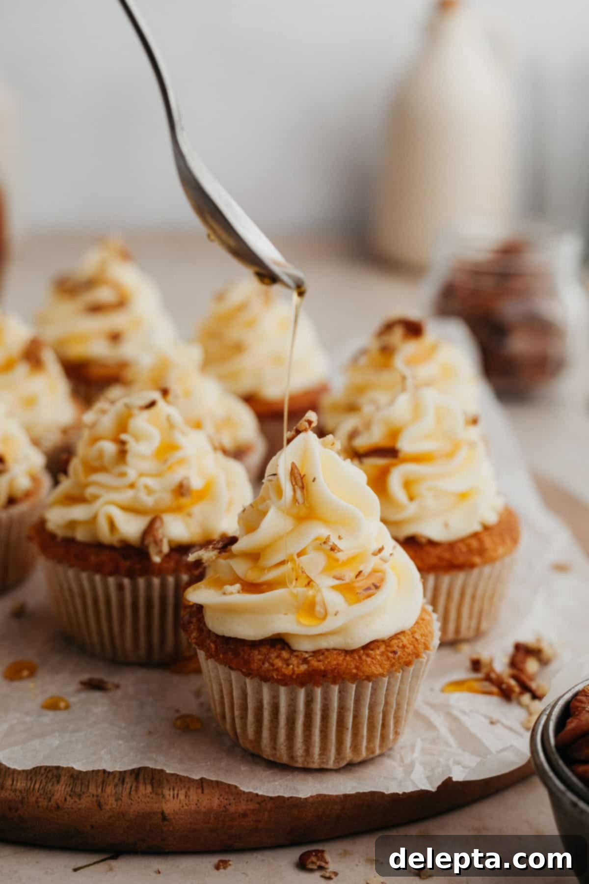 Golden Maple Delights 9 cupcakes with white frosting, the cupcake at the front is being drizzled with maple syrup and sprinkled with pecans