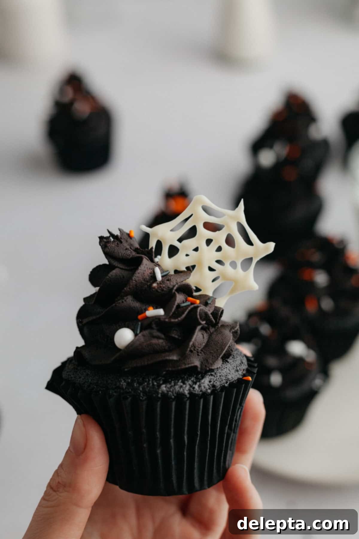Midnight Velvet Delights 9 A hand holding a black velvet cupcake with a white chocolate spiderweb on top of the frosting, perfect for Halloween.