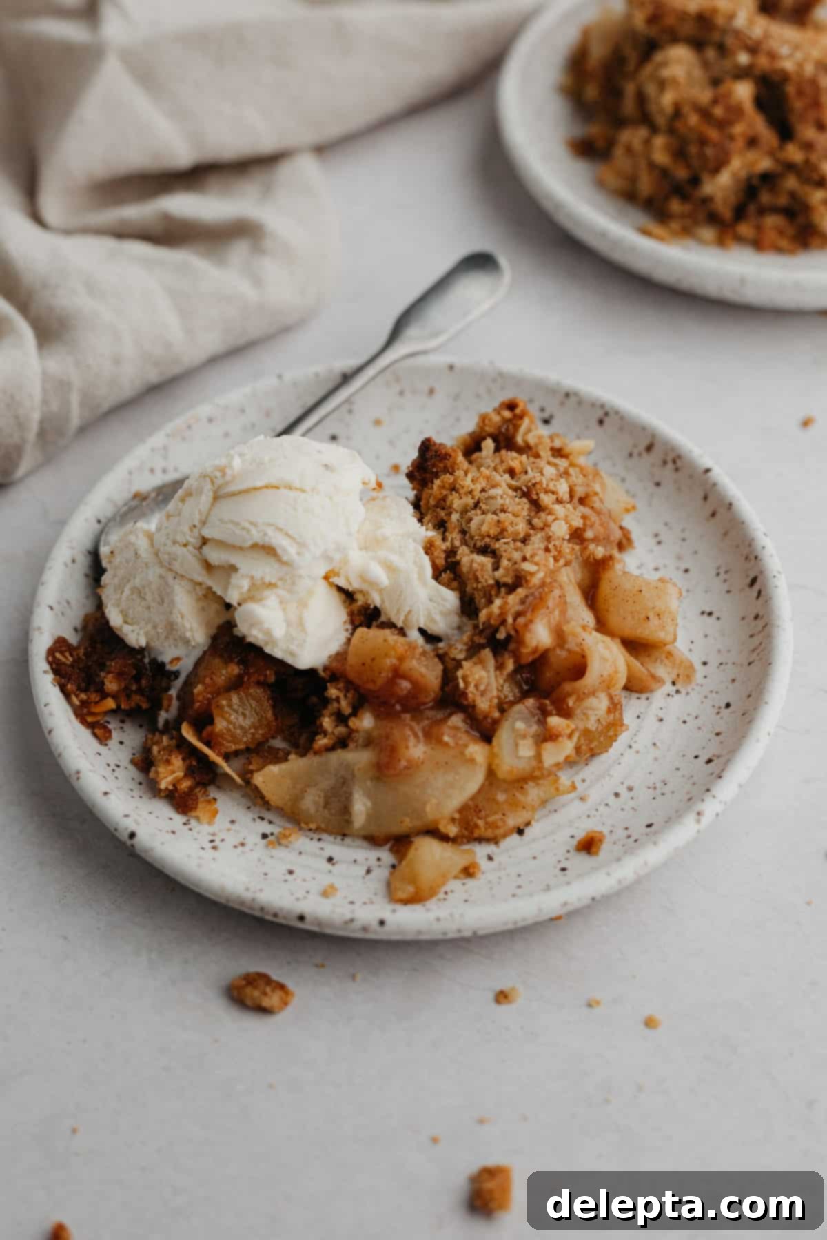 Ginger-Kissed Harvest Apple & Pear Crisp 8 A serving of warm pear and apple crisp on a white plate, garnished with a scoop of melting vanilla ice cream, sitting on a beige linen.