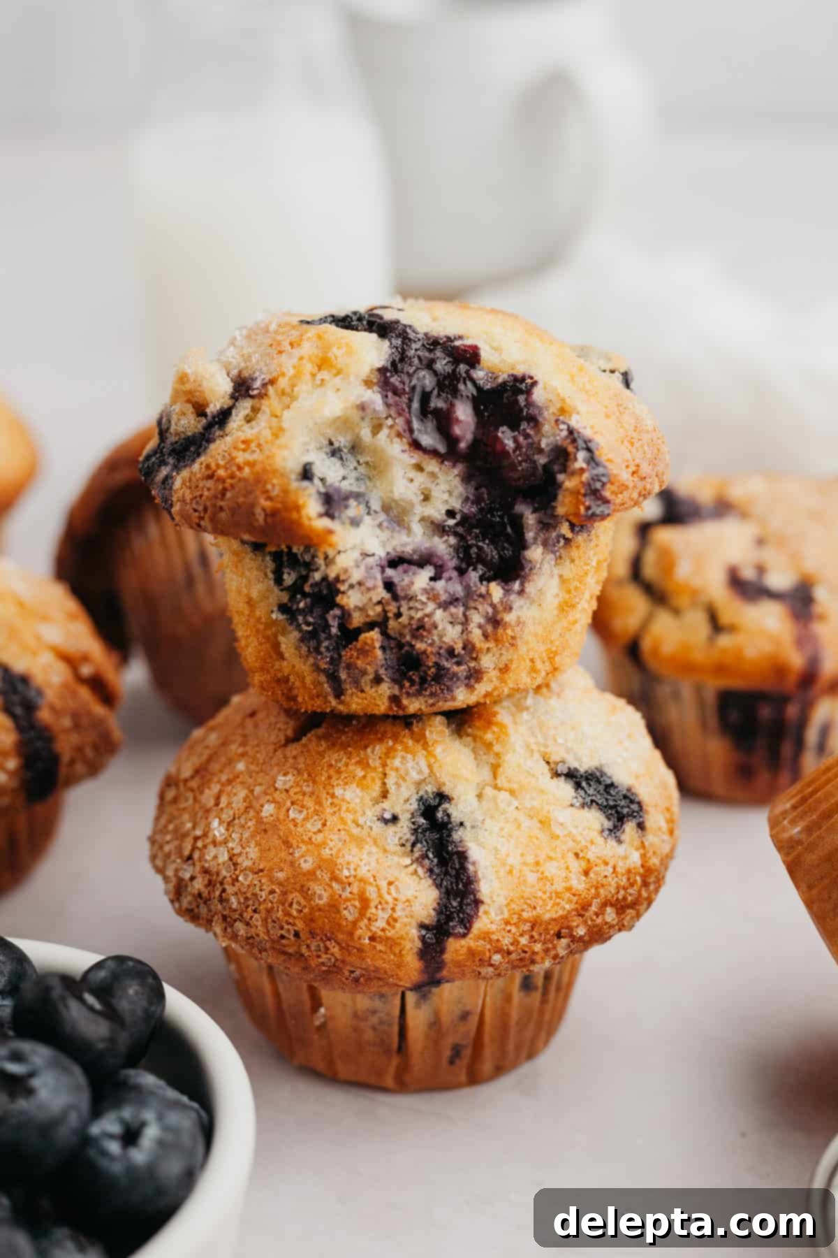 Signature Buttermilk Blueberry Muffins 6 Two perfectly baked blueberry muffins stacked on top of each other, the top one with a bite taken out, showing its moist interior.