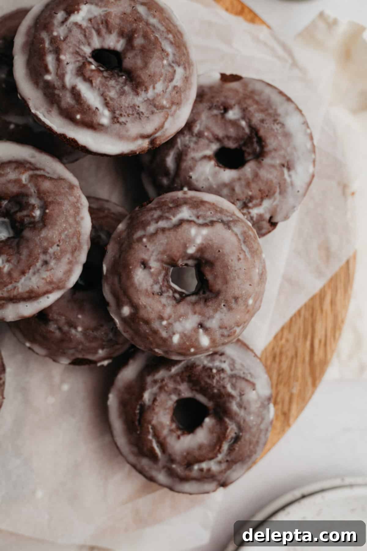 Decadent Glazed Chocolate Cake Doughnuts 2 A pile of perfectly baked chocolate cake doughnuts, glistening with vanilla glaze, arranged invitingly on parchment paper.