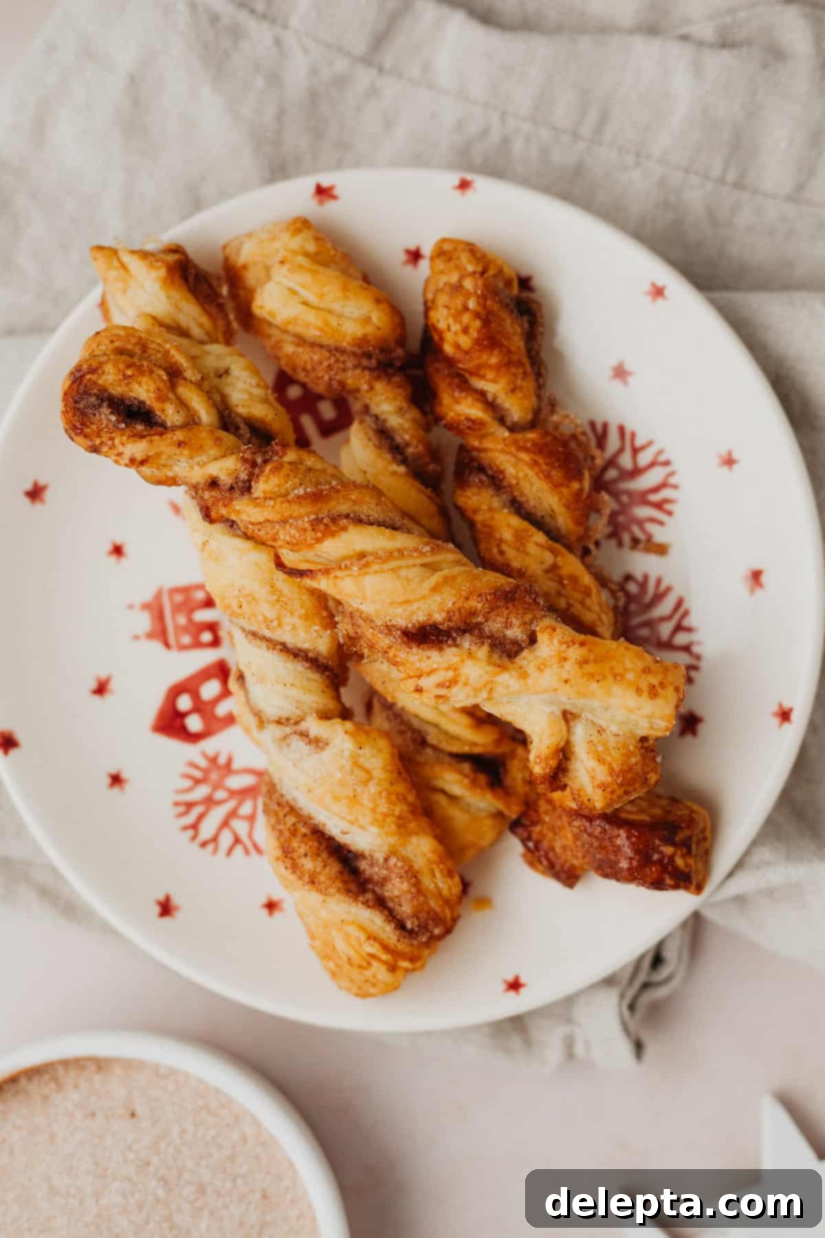 Flaky Cinnamon Twists 7 A small, festive red and white ceramic plate showcasing a stack of baked, golden-brown puff pastry cinnamon twists, glistening with melted sugar.