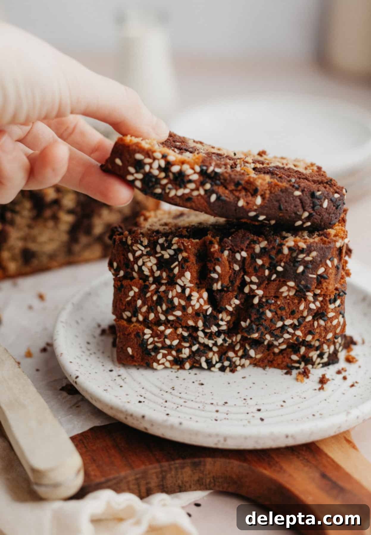 Chocolate-Tahini Swirled Banana Bread with Creamy Tang 8 A charming stack of freshly baked banana bread slices arranged on a small plate, with a hand delicately lifting one slice, inviting a taste.