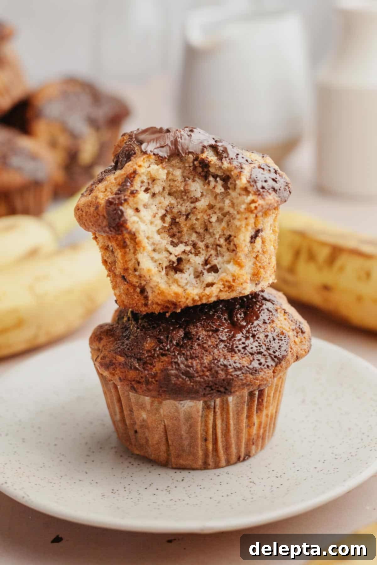 Gourmet Chocolate Chunk Banana Muffins 2 Two golden-brown chocolate chunk banana muffins stacked, one with a delectable bite taken out of it, revealing moist interior and melted chocolate.