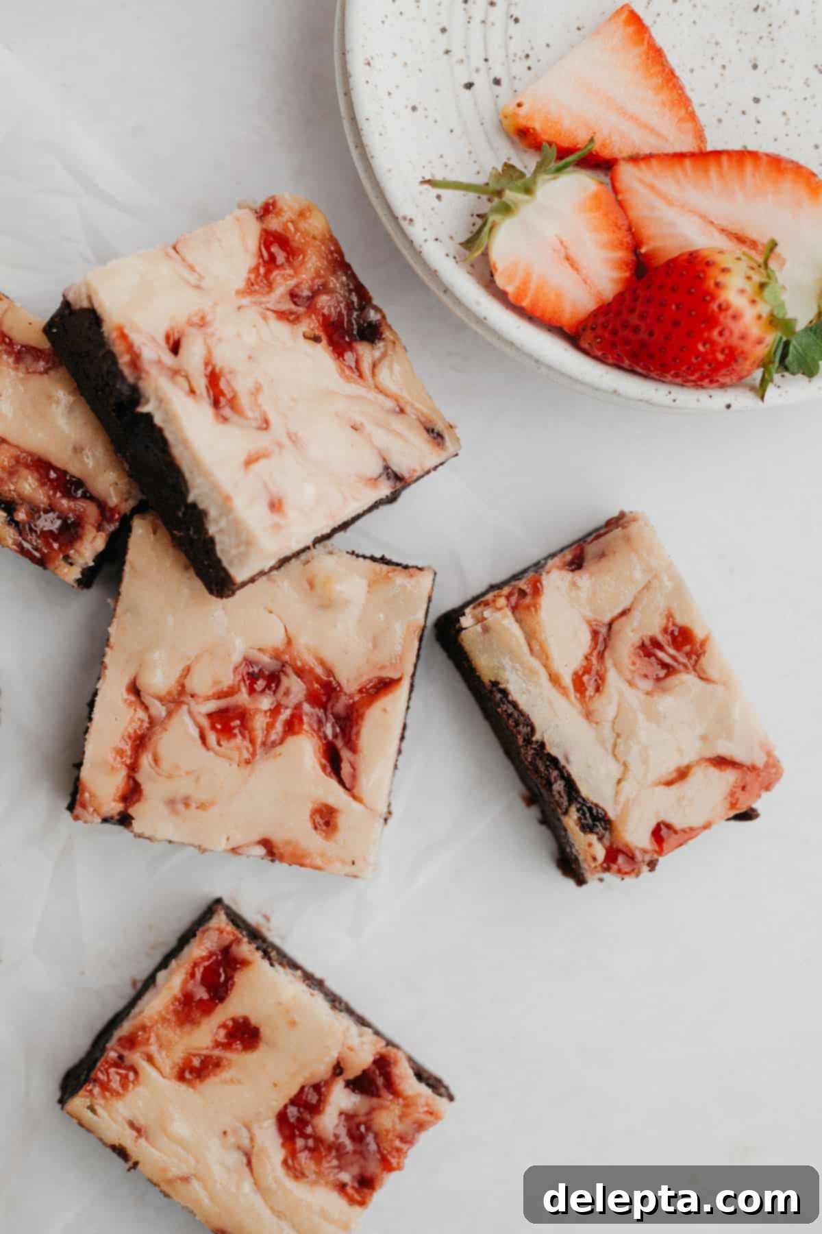 Irresistible Strawberry Cheesecake Brownies from Scratch 2 Several strawberry cheesecake brownies on parchment paper with several sliced strawberries on a grey plate.
