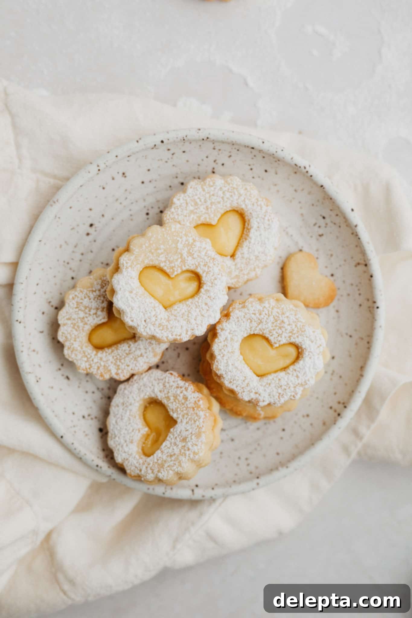 Zesty Lemon Curd Delights 2 A small, speckled beige plate filled with heart-shaped lemon curd linzer cookies, lightly dusted with confectioners' sugar. The cookies are golden brown with a vibrant yellow filling peeking through the cutout tops.