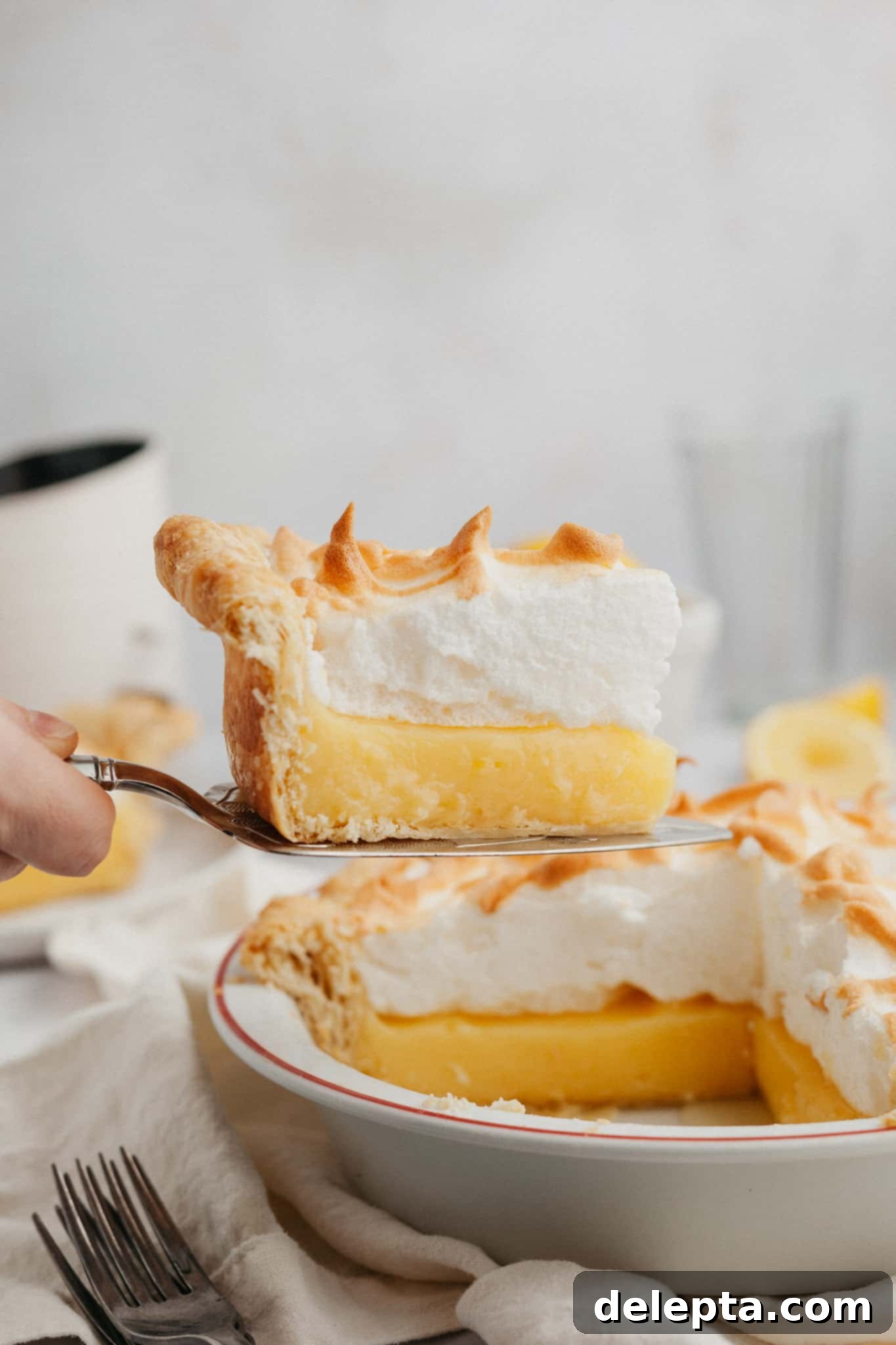 Lemon Cloud Dream 2 A beautiful slice of lemon meringue pie being lifted out with a serving knife, showcasing its distinct layers of golden meringue, bright yellow lemon curd, and flaky crust.