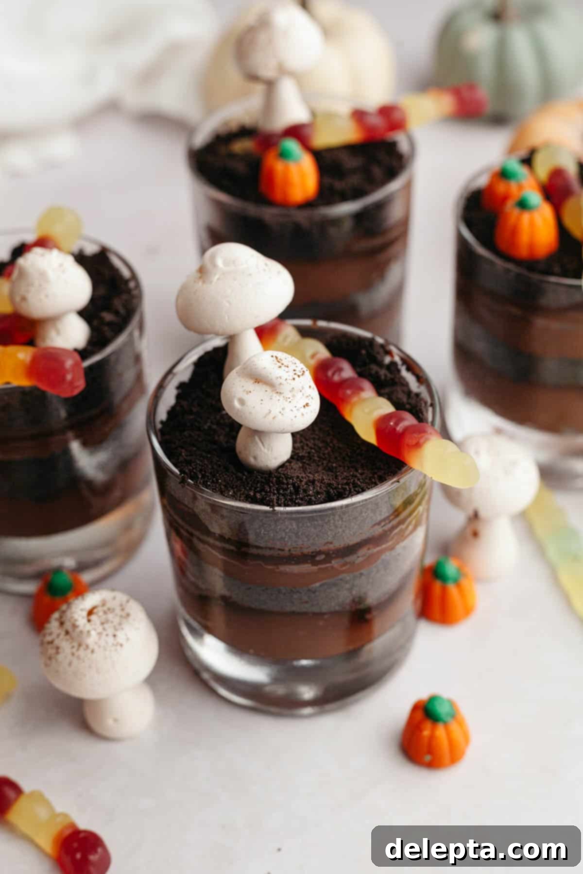 Spooky Simple Homemade Chocolate Pudding Dirt Cups 2 Glass cups with layers of chocolate pudding and Oreo cookie crumbs, decorated with meringue mushrooms and gummy worms.