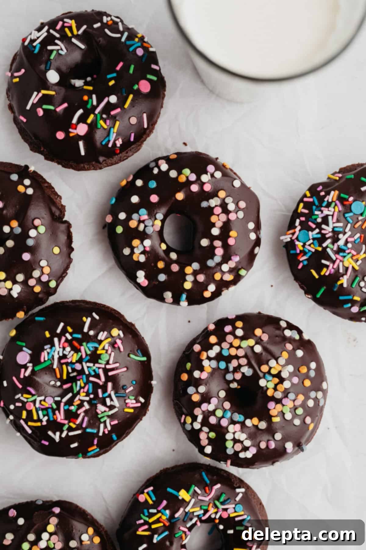 Chocolatey Sprinkled Delights 7 Freshly baked chocolate doughnuts, glistening with a rich chocolate glaze and adorned with colorful sprinkles, laid out invitingly on parchment paper.