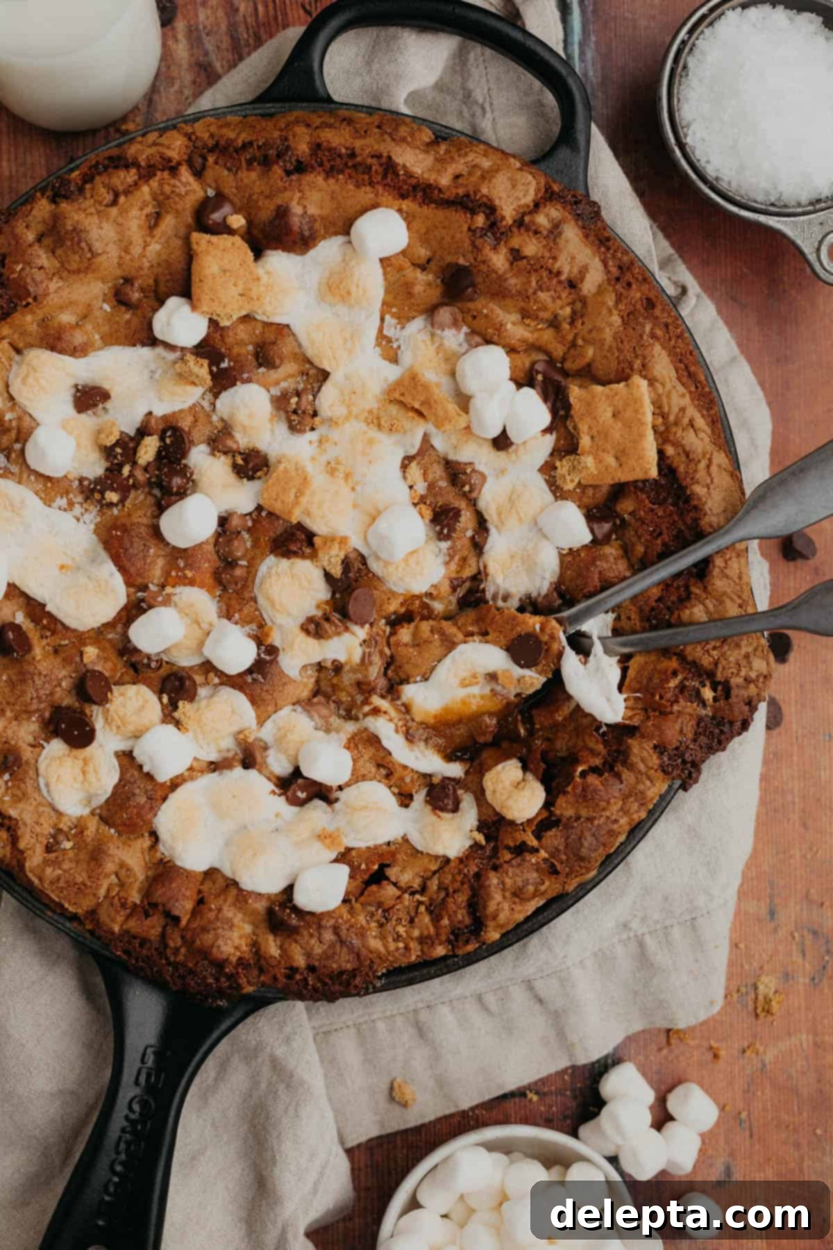 Campfire Skillet Chocolate Chip S'mores 2 A s'mores chocolate chip cookie skillet in a cast iron pan, with two silver spoons ready to scoop. The cookie is warm and gooey with melted marshmallows and chocolate puddles.