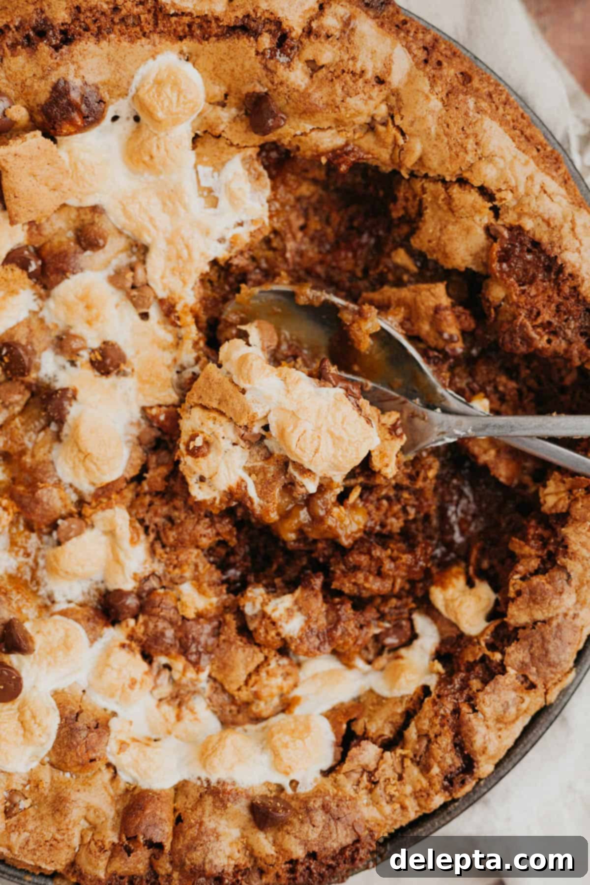 Campfire Skillet Chocolate Chip S'mores 8 A close up of a marshmallow cookie skillet, with a spoon scooping out some of the warm, gooey cookie, revealing the melted chocolate and toasted marshmallows.