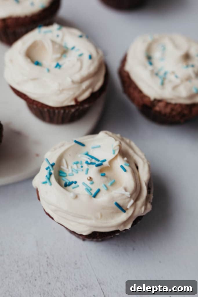 Hanukkah's Golden Glow: Chocolate, Salted Caramel & Tahini Buttercream Cupcakes 4 A close-up view of a beautifully frosted chocolate cupcake topped with a generous swirl of tahini buttercream and sparkling blue sprinkles, against a blurred background of other festive cupcakes