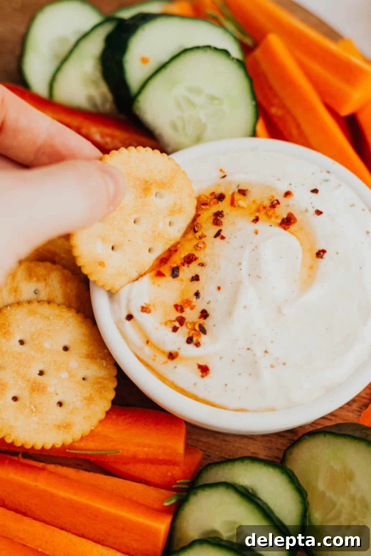 Sweet Heat Ricotta Cloud 5 A hand holding a golden cracker, dipped into a luscious white whipped ricotta dip, revealing the creamy texture and a subtle drizzle of hot honey on top. The backdrop features additional crackers and fresh vegetables, hinting at a delightful and shareable appetizer.