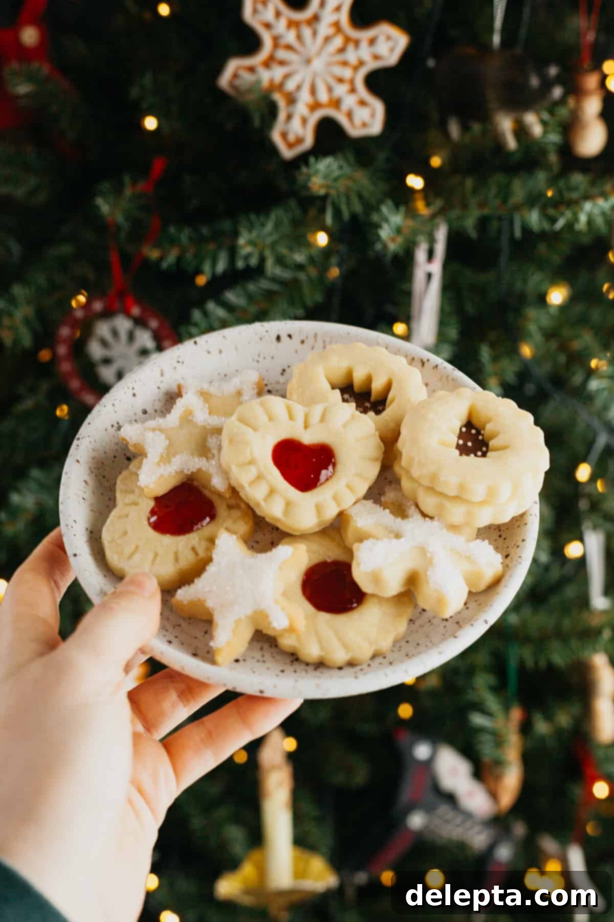 Foolproof Cutout Shortbread Cookies 7 A hand gracefully holds a small plate brimming with an assortment of beautifully decorated cutout shortbread cookies, set against the warm, blurred glow of a festive Christmas tree.