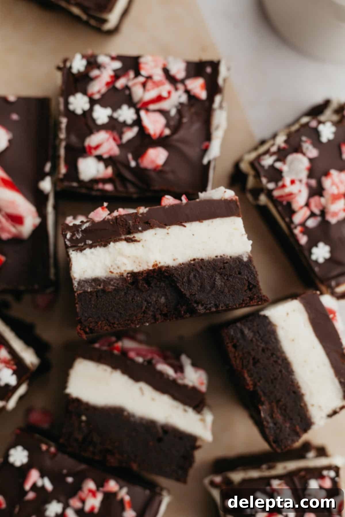 Peppermint Bark Fudge Squares 2 Beautifully layered peppermint brownies on parchment paper, showcasing the distinct layers of rich chocolate brownie, white peppermint cream cheese, and dark chocolate ganache with crushed candy canes.