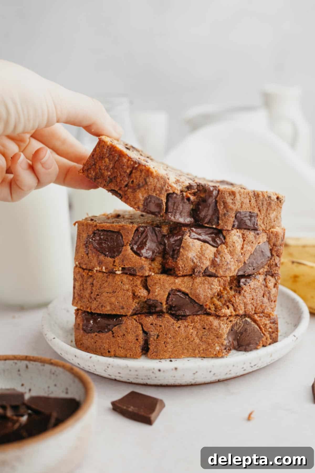 Seriously Moist Chocolate Chunk Banana Bread 10 A stack of golden brown banana bread slices on a white plate, with a hand reaching for the top slice, highlighting the inviting texture.