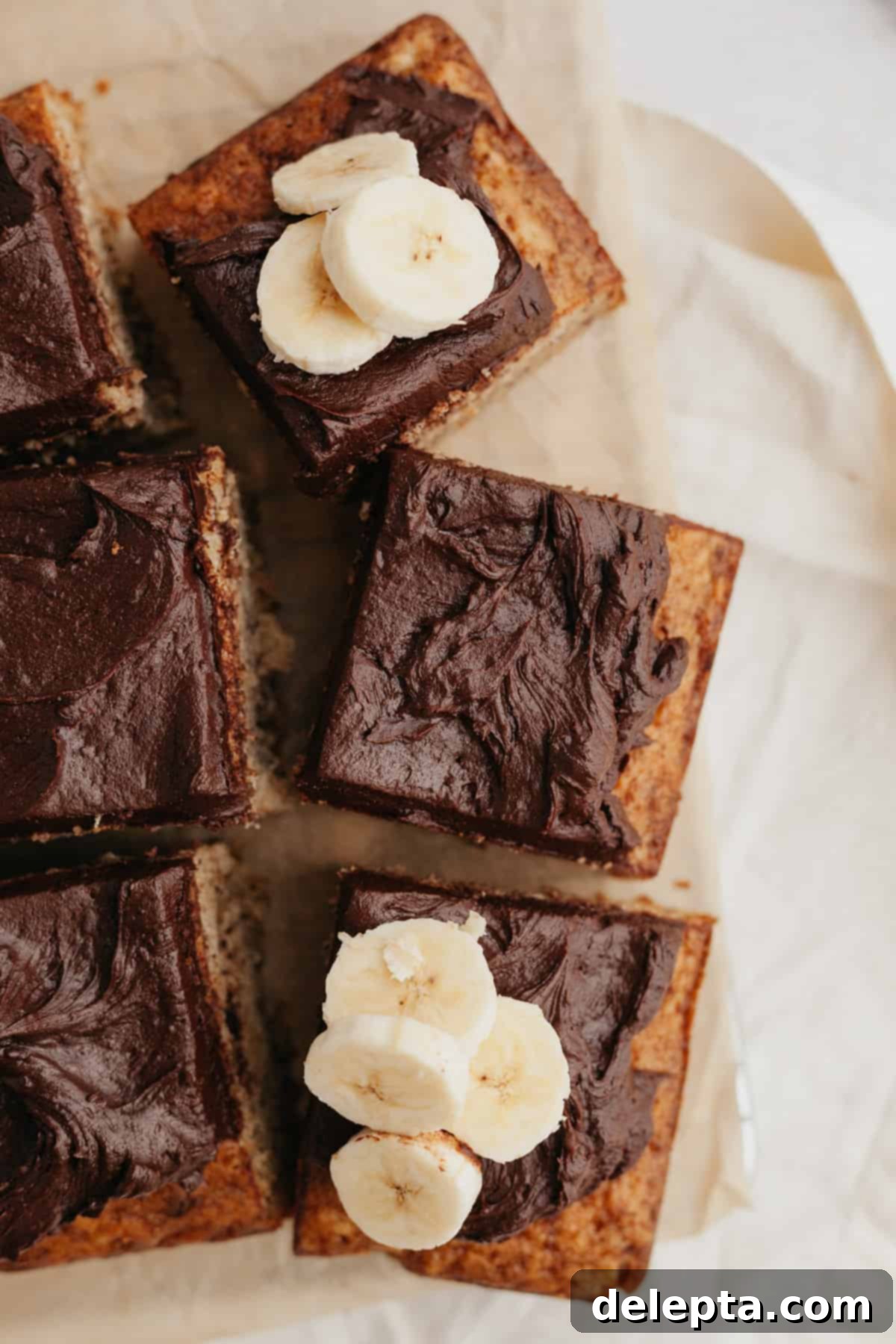 Decadent Banana Chocolate Cake 2 Squares of banana cake with rich chocolate frosting served on parchment paper, ready to be enjoyed.