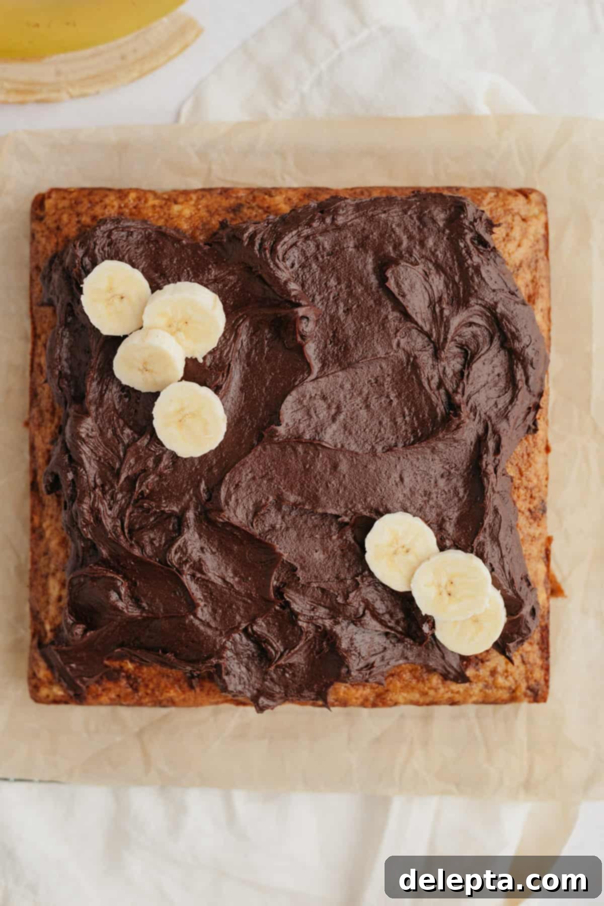 Decadent Banana Chocolate Cake 7 A beautifully frosted square banana cake adorned with fresh banana slices on top, resting on a wire rack.