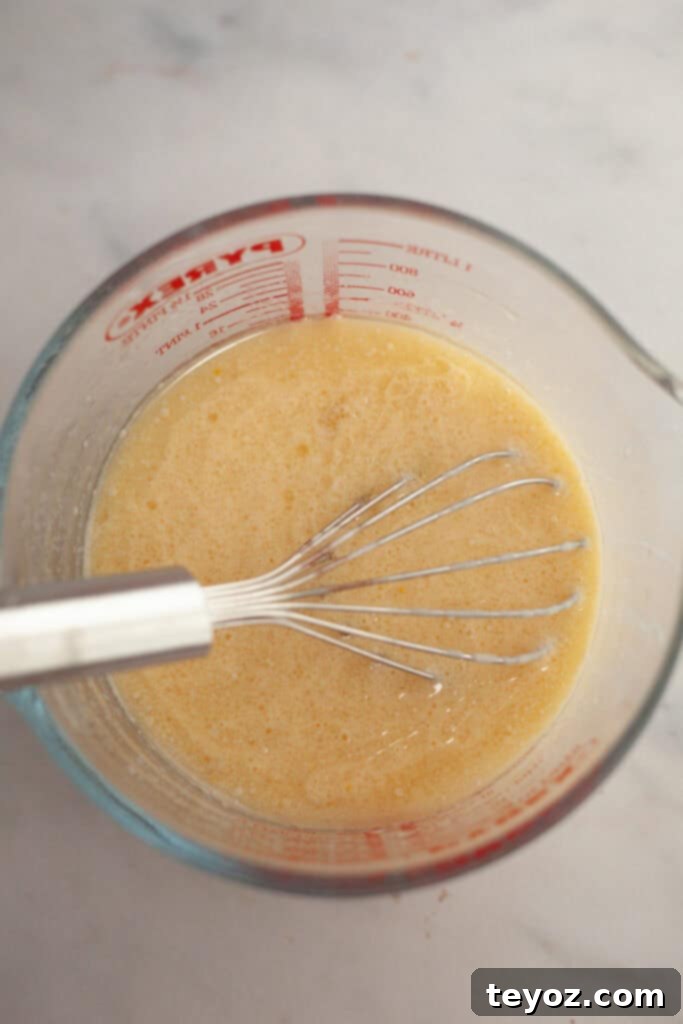 A glass jug holding a whisk and a smooth, pale yellow cake batter, illustrating the wet ingredients being thoroughly mixed before combining with the dry.