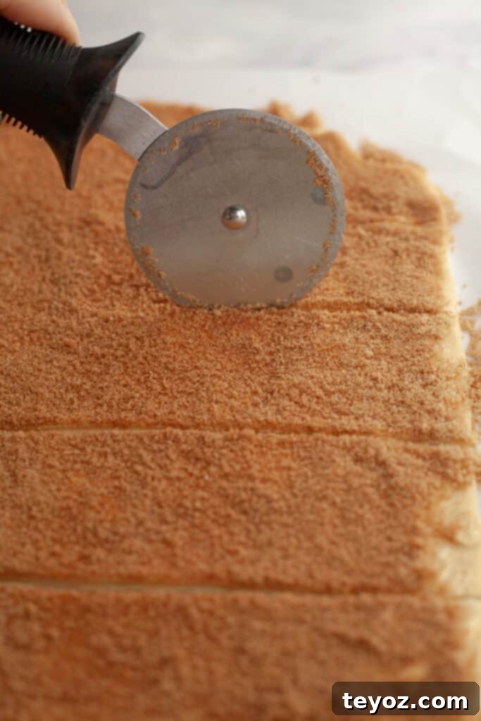 A pizza cutter slicing cinnamon roll dough into strips, demonstrating the precise cutting method.