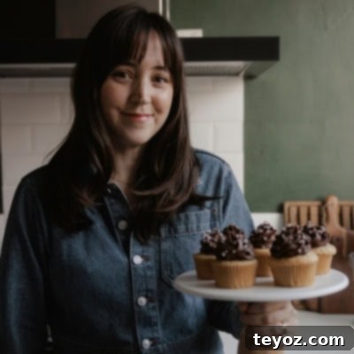Ella from Alpine Ella is holding a cake stand with cupcakes on it, smiling and inviting you to try her delicious recipes.